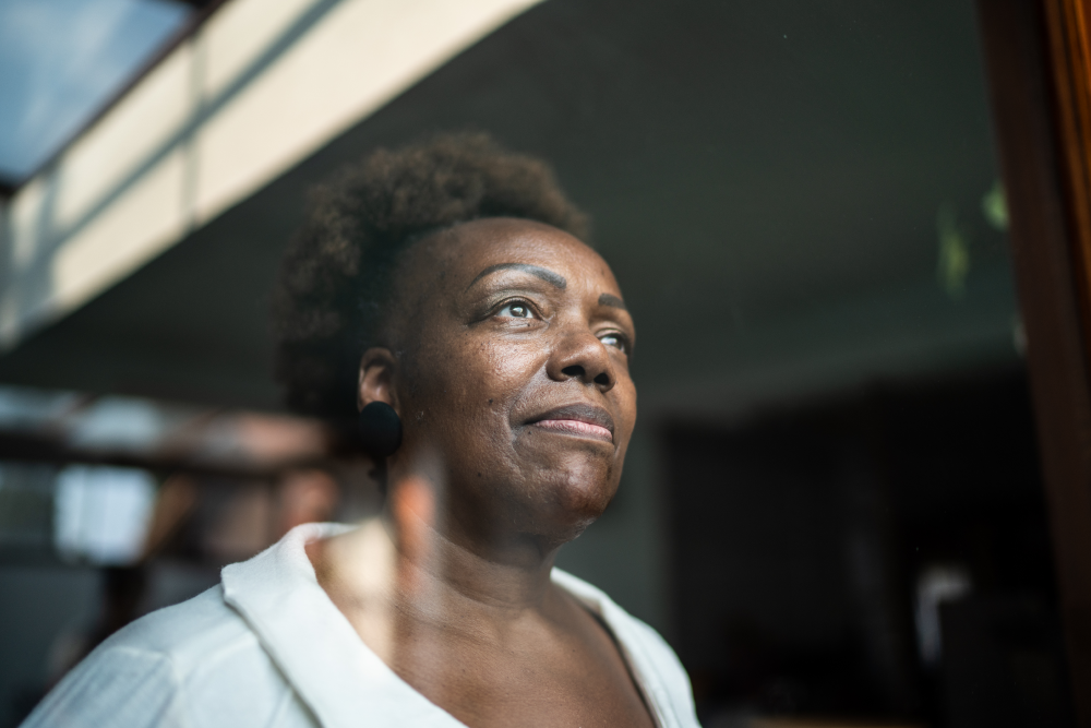 A person with a neutral expression looks out a window, wearing a white top and large earrings, with natural light illuminating their face