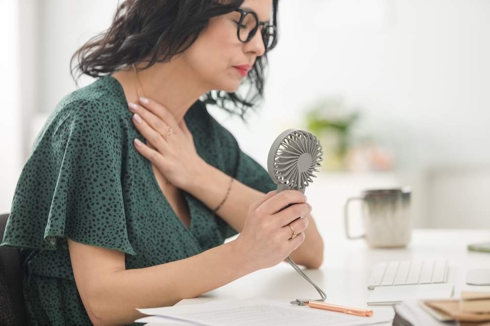 Menopausal woman wearing green dress sitting at the desk in the office, having hot flashes and using hand fan.