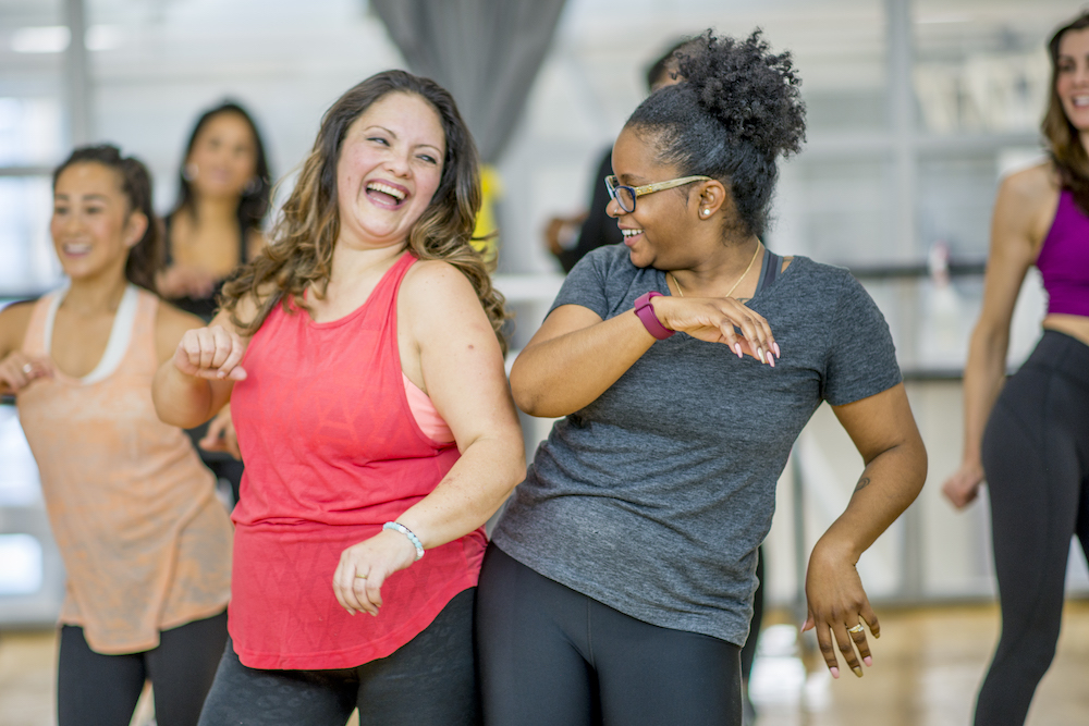 A multi-ethnic group of adult women are dancing in a fitness studio. They are wearing athletic clothes. Two women are laughing while dancing together.